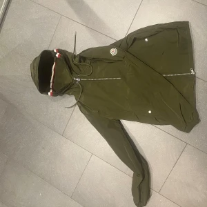 Grön vindjacka från Moncler - Snygg grön vindjacka från Moncler med huva och dragkedja. Jackan har en stilren design med Moncler-logga på bröstet och röd-vit randdetalj vid huvan. Perfekt för blåsiga dagar.