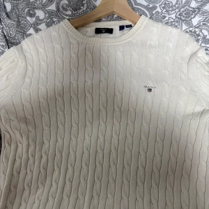 Beige stickad tröja från GANT - Säljer en stilren beige stickad tröja från GANT med kabelstickat mönster. Tröjan har en rund halsringning och är perfekt för kyligare dagar. Den har en diskret logga på bröstet.