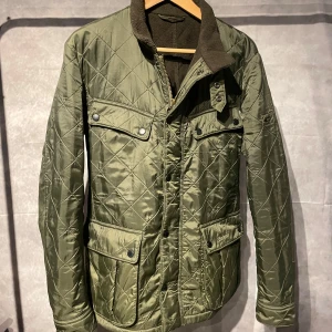  Barbour quiltad jacka   - Säljer en snygg grön quiltad jacka från Barbour. Jackan har en mjuk krage i brunt och flera praktiska fickor med knappar. Perfekt för kyligare dagar med sin stilrena design och funktionella detaljer.