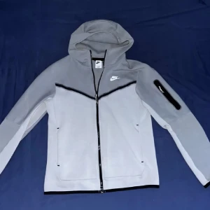 Grå tracksuit från Nike - Säljer en grå tracksuit från Nike med dragkedja och huva. Jackan har en stilren design med svart dragkedja och en liten Nike-logga på bröstet. Perfekt för en sportig look eller avslappnade dagar.
