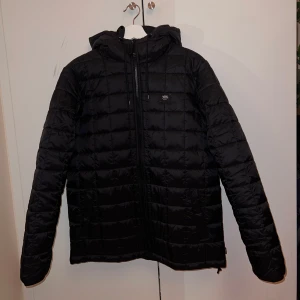 Vans Woodbridge Jacka - Säljer en svart quiltad jacka från Vans med dragkedja och huva. Jackan har en stilren design och är perfekt för kyligare dagar. Den har långa ärmar och en bekväm passform.