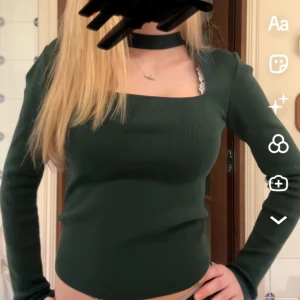 Mörkgrön ribbad topp - Snygg mörkgrön ribbad topp med lång ärm och en unik choker-detalj. Toppen har en tight passform och är perfekt för en stilren look. Den är croppad och har en fyrkantig halsringning. 