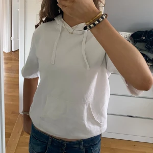 Vit hoodie med korta ärmar - Säljer en vit hoodie med korta ärmar och en praktisk magficka. Perfekt för en avslappnad stil. Den har en justerbar huva med snören. Storlek XS, bilderna är mina egna! Pris kan diskuteras 