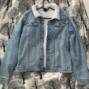 Ljusblå jeansjacka med teddyfoder - Säljer en ljusblå jeansjacka med mysigt vitt teddyfoder. Jackan har långa ärmar och knappar framtill. Perfekt för kyligare dagar!