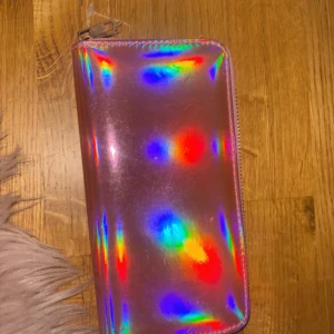 Holografisk plånbok med dragkedja - Säljer en cool holografisk plånbok i syntetmaterial med dragkedja. Plånboken har ett regnbågsskimrande yttre och rosa insida med flera fack för kort och sedlar. Perfekt för att ge lite färg till din vardag! 🌈