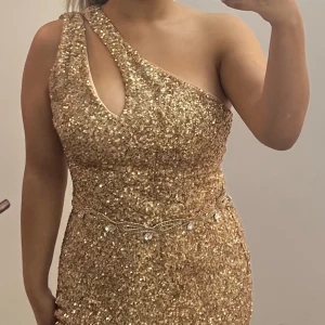 Glittrig guldklänning med slits - Snygg glittrig guldklänning med en axel och slits. Perfekt för en festlig kväll. Klänningen har en elegant design med en dekorativ kedja i midjan.