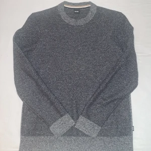 Grå stickad tröja från BOSS - Snygg grå stickad tröja från BOSS med ett diskret mönster. Tröjan har långa ärmar och en rund halsringning. Perfekt för kyligare dagar. Den är knappt använd. Den har krympt lite, så den har gått från L till M