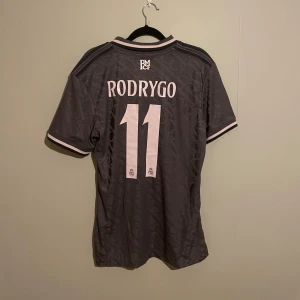 Real Madrid fotbollströja 24/25 tredjeställ - Snygg grå fotbollströja från Adidas med rosa detaljer och Real Madrid-logga. Tröjan har korta ärmar och ett stilrent mönster. Perfekt för fans av laget eller för den som gillar sportig stil.