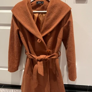 Brun kappa med huva från Deluca - Snygg brun kappa från Deluca med huva och knytbälte i midjan. Kappan har långa ärmar och knappar framtill. Dyget är gjort av polyester. Perfekt för kyliga dagar!