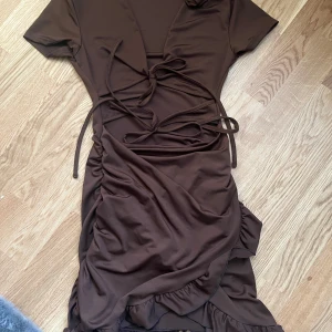 Brun klänning med volangdetaljer - Snygg brun klänning med korta ärmar och v-ringning. Den har knytning framtill och volangdetaljer längs fållen. 