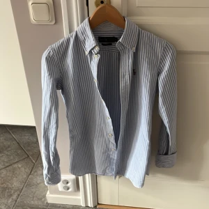 Blå Skjorta Ralph Lauren - Säljer en stilren blå- och vitrandig skjorta från Ralph Lauren i storlek XS. Skjortan har långa ärmar och knappar framtill. Den är broderad med det klassiska Ralph Lauren-logotypen på bröstet. Perfekt för en elegant look!