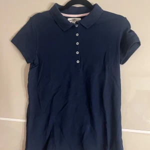 Mörkblå pikétröja från Hampton Republic - Säljer en klassisk mörkblå pikétröja från Hampton Republic i storlek XS. Tröjan har korta ärmar, krage och knappar framtill. Perfekt för en stilren och sportig look.