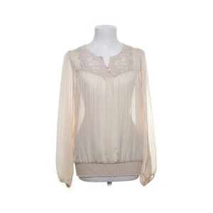 Beige blus med spetsdetaljer - Säljer en romantisk beige blus med långa ärmar och v-ringning. Blusen har fina spetsdetaljer vid halsringningen och små knappar framtill. Plisserat tyg och bred resår nedtill ger en snygg passform.