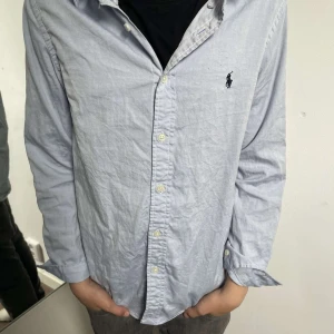 Ljusblå skjorta från Ralph Lauren - Klassisk ljusblå skjorta från Ralph Lauren med broderad logga på bröstet. Skjortan har knappar framtill och lång ärm, perfekt för en stilren look. Tillverkad i bomull för en skön känsla. en liten defekt är att de är typ 3 oerhört små hål som inte syns om man inte kollar på det as nära
