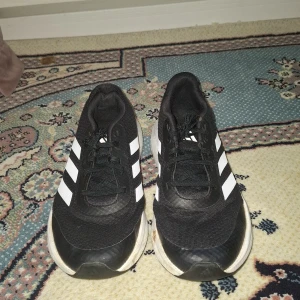 Svarta Adidas sneakers med vita ränder - Säljer ett par svarta Adidas sneakers med klassiska vita ränder på sidorna och vit sula. Skorna har snörning och är i sportig modell, perfekta för vardagsbruk eller träning. Adidaslogga på sidan och bekväm passform.