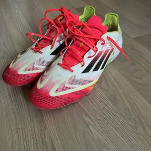 Adidas F50 Ag elite  - Säljer ett par Adidas fotbollsskor med snygg design i vitt, rött och inslag av gult. Skorna har svarta ränder på sidorna, snörning och platt sula med dobbar för bra grepp på planen. Perfekta för dig som vill sticka ut på fotbollsplanen! Påsen är även inkluderad 100% oanvänd