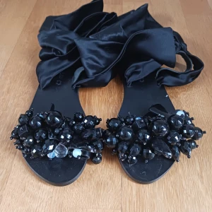 Elina Linardaki tofflor - Unika svarta sandaler från Elina Linardaki med stora svarta pärlor och stenar framtill. Skorna har breda satinband som ska gå runt benet. Handgjorda i Grekland.Strlk 36. Helt nya! Aldrig använda. Nypris ligger på 2500kr pris går att diskuteras.