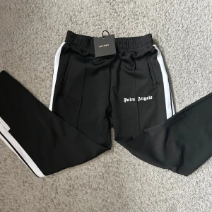 Svarta träningsbyxor från Palm Angels med vita ränder - Svarta träningsbyxor från Palm Angels med vita sidoremsor och logga tryckt på vänster ben. Byxorna har resår i midjan och raka ben. Perfekta för en sportig och trendig look.