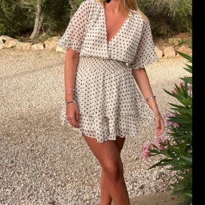 Prickig klänning  - Supersöt vit kortklänning med svarta prickar och omlottmodell. Klänningen har volangdetaljer, kort ärm och markerad midja med smock. Perfekt för sommarens alla tillfällen!