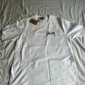 Vit t-shirt från Stüssy med tryck - Vit t-shirt från Stüssy med grönt logotryck på bröstet och stort konstnärligt tryck på ryggen. Klassisk passform och rund halsringning. Perfekt för dig som gillar streetwear och vill sticka ut med snygga detaljer.