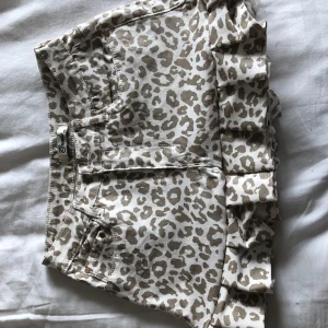 Leopardmönstrad kortkjol med volang - Säljer en beige och vit kortkjol med leopardmönster och volangdetalj nedtill. Kjolen har fickor både fram och bak samt bälteshällor. Perfekt för dig som vill sticka ut med ett trendigt mönster.