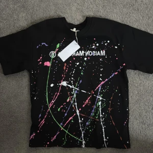 Svart t-shirt med färgstänk från Maison Margiela - Cool svart t-shirt från Maison Margiela med färgstänk i grönt, rosa, vitt och lila. Perfekt för en unik och konstnärlig stil. Kortärmad och i bomullsmaterial. Priset kan diskuteras vid snabbt köp🤩