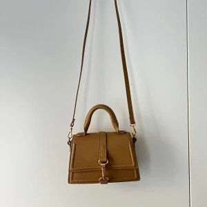 Brun axelremsväska - Säljer en stilren brun axelväska från H&M med både axelrem och handtag. Väskan har en fyrkantig form och stängs med ett snyggt spänne framtill. Perfekt för dig som vill ha en enkel men trendig accessoar. Färgen är lite ljusare i verkligheten. Priset kan diskuteras. 