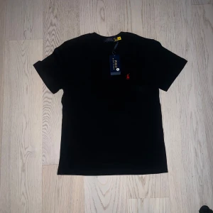 Svart t-shirt från Polo Ralph Lauren - Säljer en klassisk svart t-shirt från Polo Ralph Lauren med röd broderad logga på bröstet. T-shirten har rund halsringning och korta ärmar. Perfekt för en stilren och enkel look.