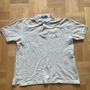Beige pikétröja från Polo Ralph Lauren - Säljer en klassisk beige pikétröja från Polo Ralph Lauren med korta ärmar och krage. Tröjan har knappar vid halsen och den ikoniska broderade loggan på bröstet. Perfekt för en stilren och avslappnad look. Står XL i nacken men passar M och L. 