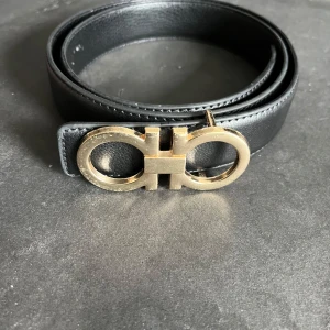 Svart bälte med guldspänne från Salvatore Ferragamo - Stilrent svart bälte i skinn med ett rundat guldspänne i Ferragamos ikoniska design. Perfekt accessoar för att lyfta din outfit med en lyxig touch. Bältet har en klassisk form och elegant finish. Skriv vid frågor