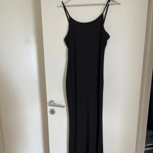 Svart långklänning med smala axelband - En stilren svart långklänning med smala axelband och figurnära passform. Perfekt för dig som vill ha en enkel men elegant look. Klänningen har en rak siluett och är ärmlös.
