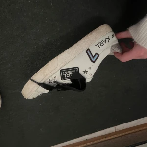 Vita sneakers Karl Lagerfeld  - Unika vita sneakers från Karl Lagerfeld med svarta sammetsband, silvriga stjärnor och nitar på tån. Sidan pryds av tryck, broderier och textdetaljer. Storlek 38.