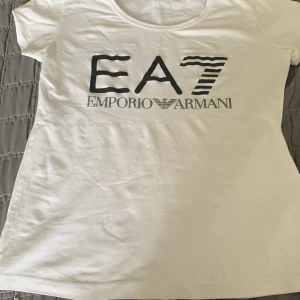 Vit t-shirt från Emporio Armani EA7 - Snygg vit t-shirt från Emporio Armani EA7 med stor svart logga framtill. Klassisk rund halsringning och korta ärmar. Perfekt för en stilren och enkel look.