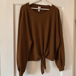 Brun långärmad topp med knyt från H&M - Säljer en brun långärmad topp från H&M med rund halsringning och knyt framtill. Toppen har en lös passform och är tillverkad i ett mjukt, strukturerat material. Perfekt för en avslappnad och trendig look.