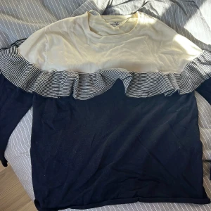 Vit och marinblå tröja med volang från Hampton Republic - Säljer en snygg tröja från Hampton Republic i storlek S. Tröjan är vit upptill och marinblå nedtill med en randig volangdetalj över bröstet och ärmarna. Perfekt för dig som gillar unika detaljer och klassisk stil.