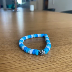 Blått pärlarmband med silverfärgad detalj - Säljer ett armband med platta pärlor i olika blå och vita nyanser. Armbandet har en silverfärgad blomformad detalj i mitten som ger en unik touch. Perfekt accessoar för att liva upp din outfit!