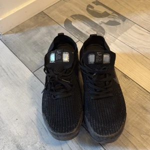 Svarta Nike Air Vapormax sneakers - Säljer ett par svarta Nike Air Vapormax sneakers med stickad ovandel och tydlig Nike-logga på sidan. Skorna har snörning och en unik, genomskinlig sula med luftbubblor för extra komfort. Perfekta för dig som gillar sportig och modern stil.