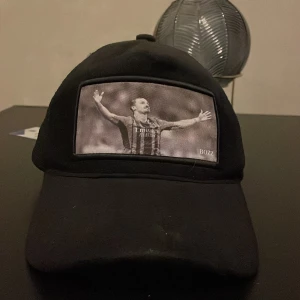 Svart keps med fotbollstryck från BOZZ - Svart keps med böjd skärm och mesh på sidorna. Framtill finns ett svartvitt tryck av en fotbollsspelare med utsträckta armar. Märket BOZZ syns på framsidan. Perfekt för dig som gillar sportig stil.