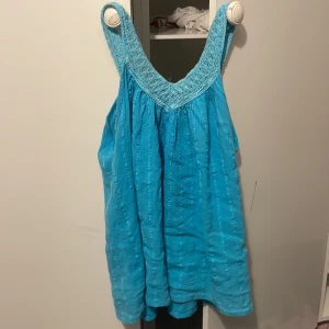 Turkos ärmlös linnetopp - Säljer en turkos ärmlös linnetopp med breda axelband och smockad överdel. Toppen är luftig och har en lös passform, perfekt för varma dagar. Materialet är lätt och känns svalt mot huden.