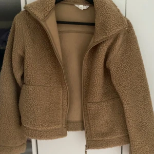 Beige teddyjacka med dragkedja - Mysig beige teddyjacka med dragkedja framtill och två stora fickor. Jackan har hög krage och är perfekt för kyligare dagar. Den har en avslappnad passform och är superenkel att matcha med olika outfits.
