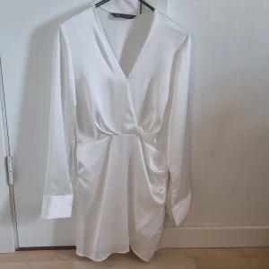 Vit omlottklänning från Zara - Säljer en elegant vit omlottklänning från Zara med lång ärm och v-ringning. Klänningen har draperad detalj i midjan och är tillverkad i ett glansigt, satinliknande material. Perfekt för dig som gillar stilrena och trendiga plagg.