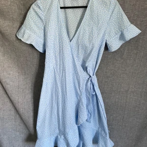 Ljusblå omlottklänning med prickigt mönster - Söt ljusblå omlottklänning med vita prickar och volangdetaljer vid ärmslut och nederkant. Klänningen har korta ärmar och knyt i midjan för en fin passform. Perfekt för varma dagar!