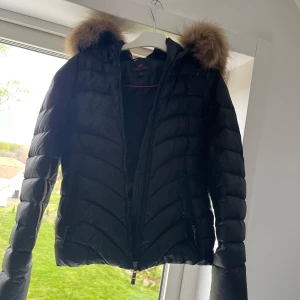 Svart dunjacka med fuskpäls från Chelsea - Säljer en svart dunjacka från Chelsea med huva och fuskpäls. Jackan har dragkedja framtill och två fickor med dragkedja. Snyggt quiltad design och långärmad modell. Perfekt för kyliga dagar.