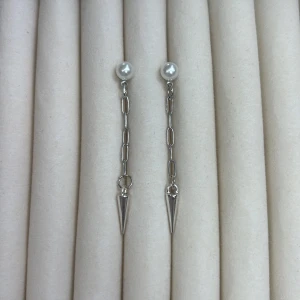Silverfärgade örhängen med pärldetalj och kedja - Snygga örhängen med vit pärla upptill, silverfärgad kedja och spetsig konformad detalj längst ner. Perfekta för att ge en edgy touch till din stil.