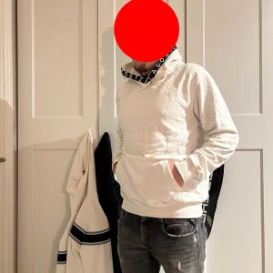 Vit hoodie från Lacoste - Säljer en stilren vit hoodie från Lacoste med stor huva och tryckt logga på kragen. Tröjan har en klassisk känguruficka framtill och ribbade muddar vid ärmslut och nederkant. Perfekt för en avslappnad look.