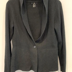 Svart stickad kavaj från H&M - Svart stickad kavaj med en knapp framtill och lång ärm. Klassisk krage och ribbad struktur, perfekt för en stilren look. Mjuk och bekväm, passar till många olika outfits.