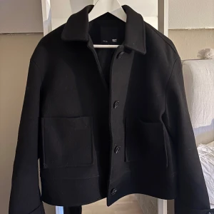 Svart kappa  - Snygg svart jacka i ull från 157 med stora framfickor. Jackan har knäppning framtill och dekorativ knappdetalj vid ärmslut. Jackan är använd ett fåtal gånger. Köpare står för frakt!