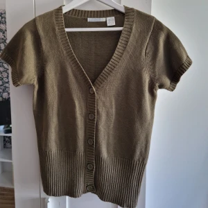 Olivgrön stickad kofta med kort ärm - Kortärmad olivgrön stickad kofta med v-ringning och knappar framtill. Ribbstickade muddar vid ärmslut och nederkant. Perfekt att slänga över en topp för en avslappnad look.