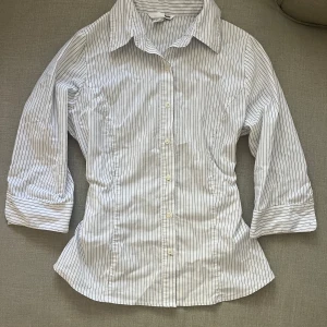 Randig figurnära skjorta - Jättefin randig skjorta som sitter åt vid midjan, och med trekvartsärm. Storlek M men kan även passa S om man vill att den sitter lösare. Finns en jätteliten fläck mellan knapparna längst ner som möjligtvis kan tas bort, annars syns det inte när skjortan är knäppt! Jättebra skick annars!