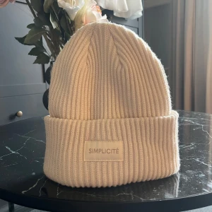 Beige ribbad mössa från HM - Snygg beige ribbstickad mössa från hm med uppvikt kant och diskret logotyp framtill. Perfekt accessoar för kyliga dagar och enkel att matcha med olika outfits.